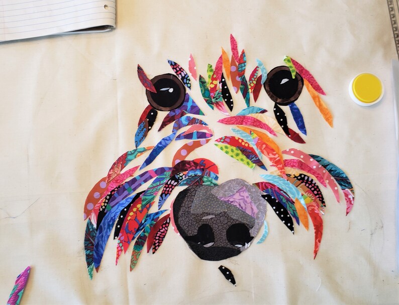 Pode incluir: Uma obra de arte de tecido colorida que representa o rosto de um c&atilde;o. A pelagem do c&atilde;o &eacute; feita de v&aacute;rios peda&ccedil;os de tecido coloridos, e os olhos s&atilde;o pretos com pupilas brancas. O nariz &eacute; um tecido cinza escuro com um padr&atilde;o floral.
