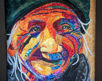 Pode incluir: Um retrato colorido de uma pessoa sorrindo, feito de retalhos de tecido. A pessoa está usando um chapéu e tem cabelo branco. O retrato está emoldurado em uma moldura preta.