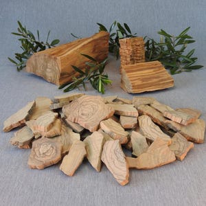 Puede incluir: Una colección de piezas de madera de olivo, incluyendo bloques más grandes y rodajas más pequeñas e irregulares. La madera muestra un color cálido y natural con patrones de vetas distintivos. Ramas verdes de olivo están dispuestas en el fondo.