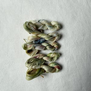 Può includere: Cinque piccole matassine di filo da ricamo ritorte disposte in una pila verticale. Il filo presenta una tavolozza di colori variegati composta da verde salvia tenue, oliva pallido, crema e lievi accenni di rosa polvere e azzurro.