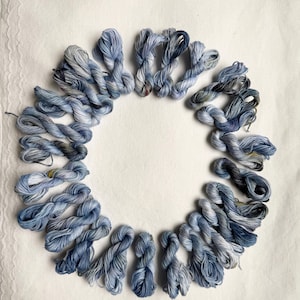 Può includere: Disposizione circolare di matassine di filo da ricamo blu e grigio. Il filo è disposto su una superficie bianca con un bordo in pizzo. Le matassine variano nelle tonalità di blu, da chiaro a scuro, creando un effetto sfumato.