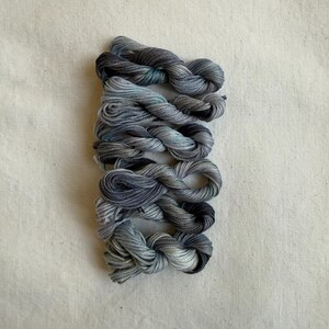 Può includere: Una collezione di sei matassine di filo da ricamo tinto a mano in tonalità di grigio, blu e nero. Il filo è disposto verticalmente su uno sfondo di tessuto neutro. Ogni matassina è ben avvolta e mostra una sfumatura di colore.