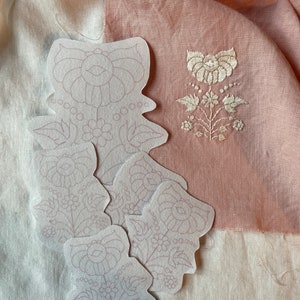 May include: Five white fabric embroidery patterns with a floral design. The patterns are layered on top of each other. The patterns are on a white fabric background.