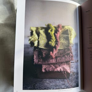 May include: A stack of colorful fabric swatches in shades of green and pink, with frayed edges and textured surfaces. A pink and red yarn is draped over the top. The image is from a cookbook, with the text "ALONG THE PATHS, WILD PLANTS" at the bottom.
