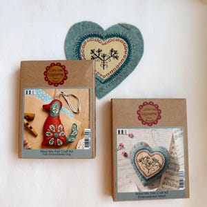 Pode incluir: Três kits de artesanato e um coração de feltro sobre uma superfície branca. Os kits estão em caixas castanhas com o texto "Corinne Lapierre" e "Wool Mix Felt Craft Kit". Um kit mostra um cão de feltro vermelho, outro um coração azul.