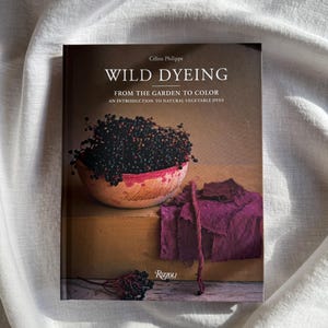 May include: A book titled "WILD DYEING" by C&eacute;line Philippe, with the subtitle "FROM THE GARDEN TO COLOR." The cover features a bowl of dark berries, a swatch of purple fabric, and the Rizzoli logo. The book is on a wooden surface.