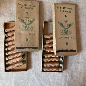 May include: Two rectangular cardboard boxes containing spools of light brown sewing thread. The boxes are labeled with the text SOIE NATURELLE A COUDRE, AU PHENIX, and S.A. LA SOIE, featuring a green graphic illustration of a phoenix bird.