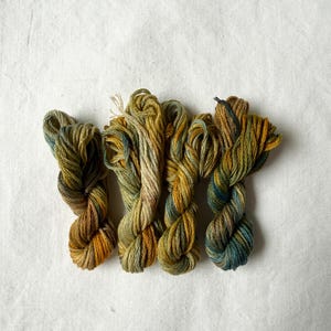 Può includere: Quattro matasse di filo da ricamo tinto a mano disposte fianco a fianco. I fili presentano una tavolozza di colori variegati composta da verde oliva terroso, giallo senape e blu ottanio profondo, con sottili variazioni tonali.