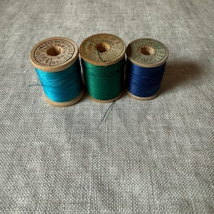 May include: Three vintage wooden spools of thread in shades of turquoise, emerald green, and navy blue. Each spool features a circular paper label on top with printed text including the words Waxed Mercerized Cotton and Belding.