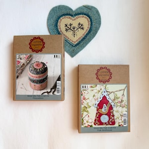 Op de afbeelding: Twee knutselsets en een vilten hart op een witte achtergrond. De sets zitten in bruine dozen met de tekst "Corinne Lapierre" en "Wool Mix Felt Craft Kit". Eén set is voor een speldenkussen, de andere voor een volks vogelhuisje. Het hart is blauw met geborduurde details.