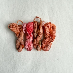 Può includere: Quattro matasse di filo da ricamo disposte in fila. La prima è di un arancione tenue, la seconda presenta una vivace sfumatura di rosa e rosso, la terza è di un arancione brunastro e la quarta è di un arancione brillante. Ogni matassa è attorcigliata.