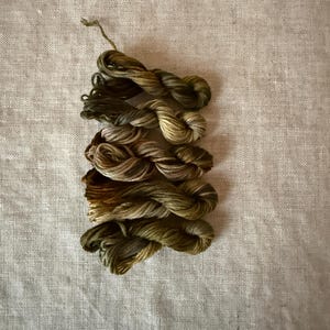 May include: A collection of five small, twisted skeins of embroidery thread arranged in a vertical stack. The threads feature variegated color palettes in shades of olive green, moss green, golden brown, and muted tan with subtle tonal shifts.