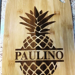 May include: A brown bamboo cutting board with a pineapple design and the name "PAULINO" engraved in the center.