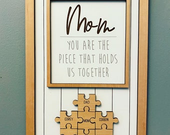 Puede incluir: Un marco de madera con fondo blanco y el texto "Mom You Are The Piece That Holds Us Together". Debajo del texto hay un rompecabezas de madera con los nombres "Dad", "Corey", "Mom", "Corbin", "Laney", "Nixon" y "Eli" en las piezas.
