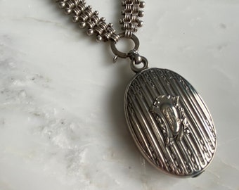 May include: A silver locket necklace with a chain made of linked circles. The locket is oval shaped and has a detailed engraved design with a floral motif.