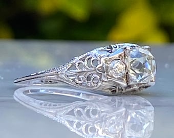 Könnte beinhalten: Ein Vintage-Verlobungsring mit filigranem Design, einem großen, klaren Mittelstück und einem kleineren Diamanten als Akzent. Der Ring ist aus weißem Metall gefertigt und hat ein zartes, filigranes Design.