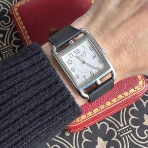 Könnte beinhalten: Eine rechteckige silberne Armbanduhr mit weißem Zifferblatt, schwarzem Lederarmband und Datumsanzeige. Die Uhr hat große Ziffern und wird am Handgelenk getragen. Die Uhr befindet sich neben einem roten Etui.