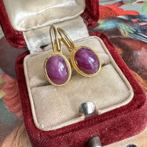 May include: A pair of gold earrings with oval-shaped purple gemstones. The earrings are in a red velvet jewelry box.