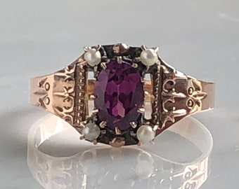 Könnte beinhalten: Ein Vintage-Ring mit einem tiefvioletten Edelstein, der von vier kleinen Perlen umgeben ist. Der Ring ist aus Gold gefertigt und hat ein filigranes Design.