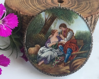 Könnte beinhalten: Ein runder Anhänger mit silbernem Rahmen, der eine gemalte Szene eines Paares in einer Waldlandschaft zeigt. Die Frau trägt ein lila-weißes Kleid, der Mann eine blaue Hose und ein rot-weißes Hemd. Die Szene ist realistisch gemalt mit Details wie Bäumen, Blumen und einem Schaf.