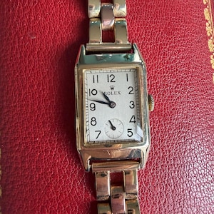 May include: A gold Rolex wristwatch with a rectangular face and a gold strap. The watch face is white with black numerals and hands. The Rolex logo is visible above the centre of the face.