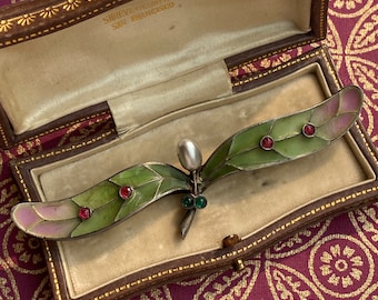 May include: A delicate, silver dragonfly brooch with green and red enamel wings and a pearl accent. The brooch is displayed in a brown velvet box with the inscription "Shreve & Company San Francisco".