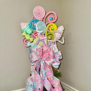 May include: A whimsical candy-themed tree topper featuring colorful lollipops, cupcakes, and swirls. The design includes a large pink bow with ribbons printed with ice cream cones and the word "HO". The tree topper is set in a black pot.
