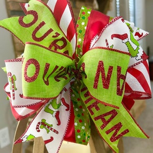 Pode incluir: Um laço de Natal festivo com fitas verdes e vermelhas. O laço tem as palavras "Sure Mean" em glitter vermelho em fitas verdes. Outras fitas apresentam desenhos de bengalas doces e do Grinch. As fitas listradas em vermelho e branco completam o tema festivo.