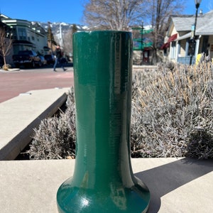 May include: A dark green ceramic vase with a flared base. The vase is sitting on a concrete surface.