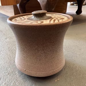 Peut inclure: Un pot en céramique marron avec un couvercle. Le pot a une surface texturée et une forme arrondie, avec un couvercle orné d'un motif tourbillonnant. Le pot est probablement utilisé pour le stockage et a un aspect rustique et fait à la main.