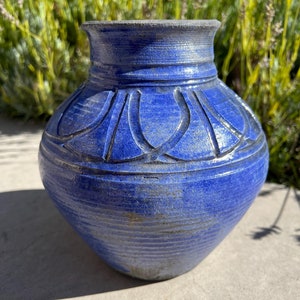 Può includere: Un vaso in ceramica blu con una superficie testurizzata e un design intagliato intorno al centro. Il vaso ha un corpo arrotondato e un'apertura leggermente svasata.