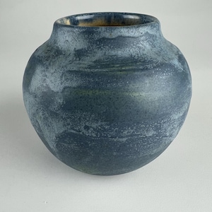 May include: A round ceramic vase with a wide body and a narrow neck opening. The exterior features a mottled, textured finish in shades of dark blue and slate grey, with a smooth, glossy golden-brown glaze visible on the interior rim of the vessel.
