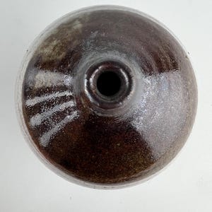 May include: A dark brown ceramic bottle, viewed from above, with a glossy finish. The bottle has a rounded shape, a narrow neck, and a small opening. The surface shows subtle colour and texture variations. The bottle is likely handmade.