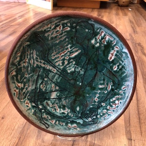May include: A large, round ceramic plate with a green and white glaze. The plate features a black silhouette of a figure in a dancing pose.
