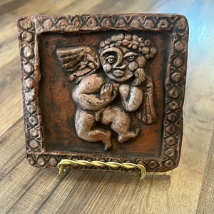 May include: A rectangular, brown ceramic tile featuring a relief sculpture of a cherub with wings. The cherub is in a seated position, with its head resting on its hand. The tile has a decorative border and is displayed on a gold wire stand.
