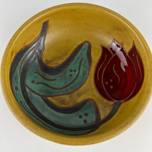 May include: A round ceramic bowl with a mustard yellow base color. The interior features a hand-painted design of a dark red tulip flower with a long, curved green stem and two large green leaves, all outlined in a dark brown or black color.