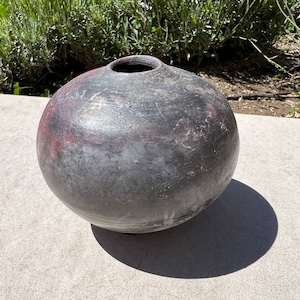 Könnte beinhalten: Eine runde, schwarze Keramikvase mit leicht strukturierter Oberfläche. Die Vase hat einen dezenten rotbraunen Farbton und eine breite Öffnung oben.