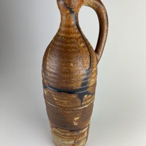 Puede incluir: Una jarra de cerámica artesanal con asa. La jarra presenta una combinación de colores marrón y beige con detalles en marrón oscuro y negro. La cerámica tiene una superficie texturizada y una forma orgánica única. Esta pieza decorativa es adecuada para la decoración del hogar.