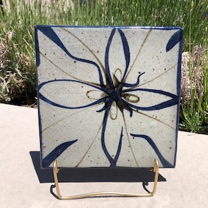 Puede incluir: Un azulejo de cerámica cuadrado con un diseño floral azul y blanco. El azulejo está en un soporte de metal dorado.