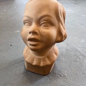 May include: A vintage bisque doll head with closed eyes and a ruffled collar. The unpainted ceramic head has a smooth texture and a light tan color. The doll head is a decorative item.