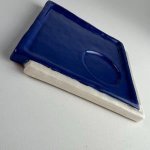 Könnte beinhalten: Ein Keramiktablett mit tiefblauem Innenraum und weißer, rechteckiger Basis. Das Tablett hat eine rautenförmige Form mit einer kreisförmigen Vertiefung. Die glänzende Oberfläche reflektiert das Licht.