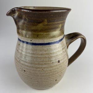 Può includere: Brocca in ceramica fatta a mano con beccuccio e manico. La brocca presenta un corpo beige maculato, un bordo e un manico marrone scuro. Una sottile striscia blu circonda il centro. Adatta per servire bevande.