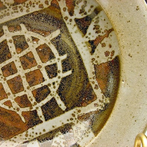 Könnte beinhalten: Ein Keramikteller mit weißer Glasur und einem braunen und orangen Design. Das Design zeigt ein Gittermuster mit einem Kreis in der Mitte.