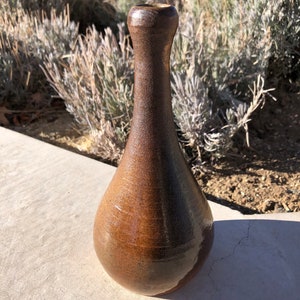 Puede incluir: Un jarrón de cerámica marrón con fondo redondeado y cuello estrecho. El jarrón tiene un esmalte moteado y un acabado brillante.