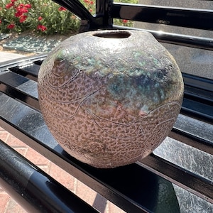Puede incluir: Un jarrón redondo de cerámica texturizado con un esmalte verde y marrón. El jarrón tiene una pequeña abertura en la parte superior y está sentado en un banco de metal negro.