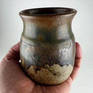 May include: A small, rounded ceramic cup featuring a reactive glaze with blended tones of dark brown, muted green, and a textured, speckled beige base. The rim is smooth and slightly flared, transitioning into a deep, earthy color palette.