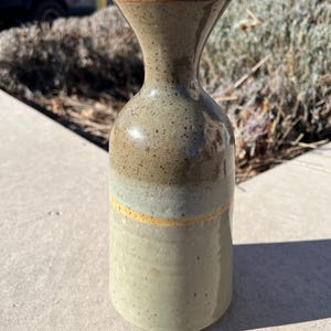 以下が含まれることがあります： 手作りの陶器の花瓶。土を思わせる色合いで、斑点模様が特徴。上部はフレア状で、円筒形の胴体には茶色、青灰色、緑色の帯がある。細い黄色の帯がデザインを強調している。