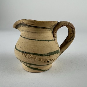 May include: A small, handcrafted ceramic pitcher with a rounded body and a handle. The pitcher is beige with dark green horizontal stripes and the words "Silver Serling" are embossed on the side. The spout is curved.