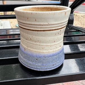 May include: A handmade ceramic vase with a wide opening. The vase features a light beige upper section, a brown band, and a blue-grey base. The exterior has a speckled texture, and the interior is a darker brown.