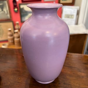 May include: A lavender-colored ceramic vase with a rounded body and a slightly flared rim. The vase has a smooth, matte finish and sits on a wooden surface. The background features framed artwork.